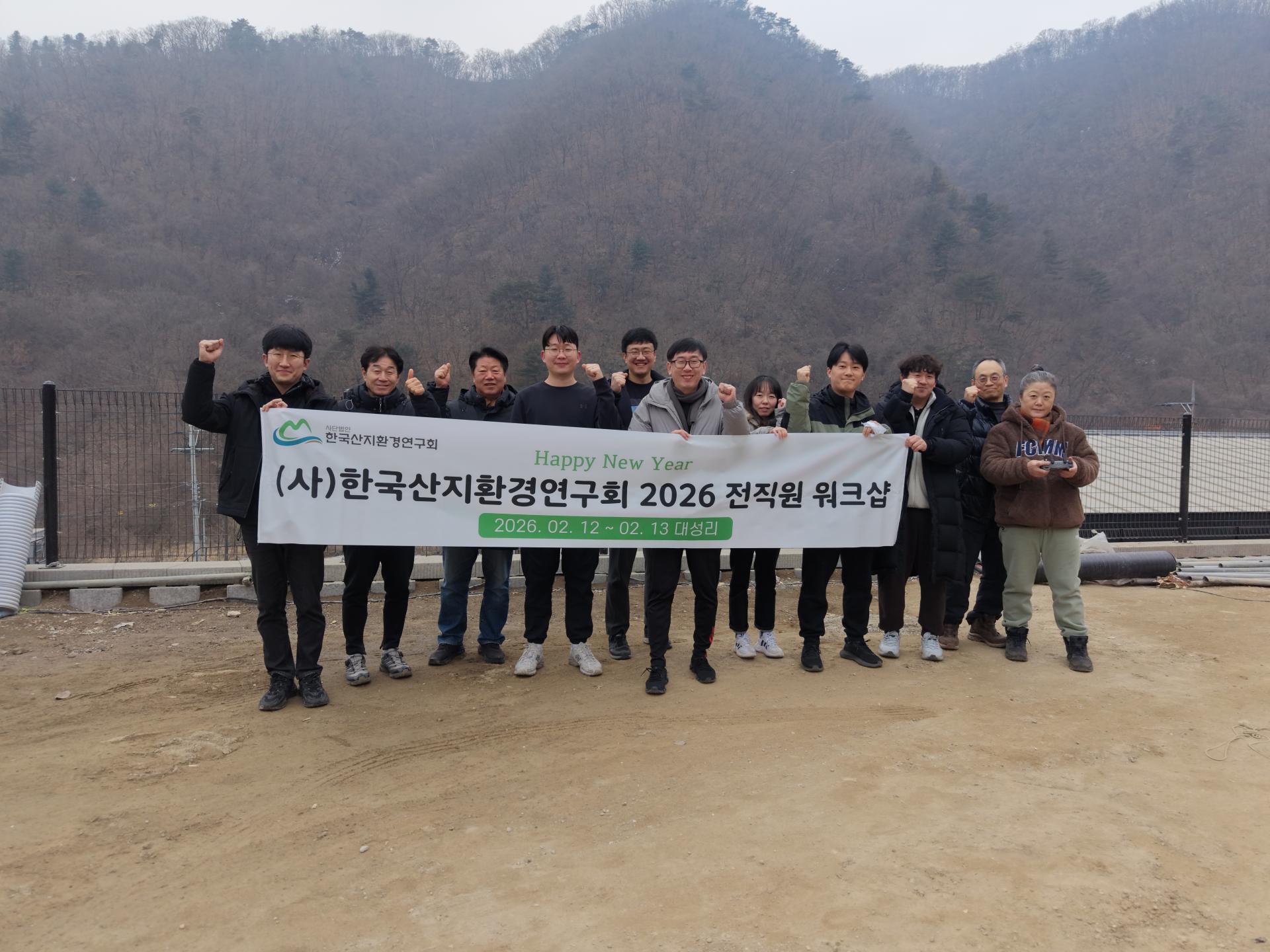 산을 배경으로 '2026 전직원 워크샵' 현수막을 들고 단체로 기념하는 직원들 (2026년 2월 12~13일)